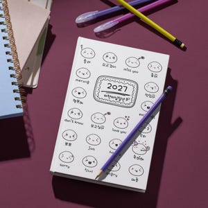 Peut inclure: Un agenda blanc 2027 orné d'une grille de visages de dessins animés aux expressions variées, accompagnés de textes en anglais et en coréen. Un crayon violet est posé en diagonale sur la couverture, avec d'autres stylos à proximité.