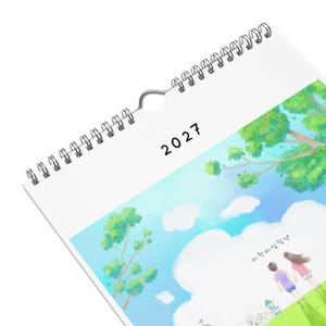 Puede incluir: Un calendario de pared para el año 2027 con una espiral de metal plateado y un orificio central para colgar. La portada presenta una ilustración digital de un cielo azul con nubes blancas, árboles verdes y dos figuras sobre una colina.