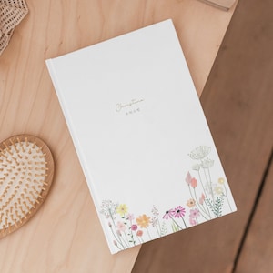 May include: A white journal with the name "Christina" written in gold lettering and a floral design on the bottom. The journal is on a wooden surface.