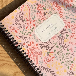 May include: A pink spiral bound notebook with a floral pattern and the text "내가 사랑하는, that I love" in a white oval.