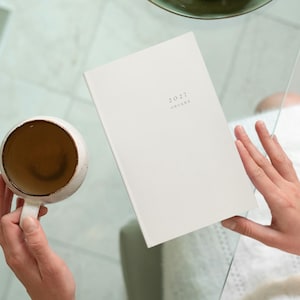 Peut inclure: Un agenda rectangulaire blanc avec le texte 2027 imprimé sur la couverture avant, placé à côté d'une tasse en céramique ronde contenant un liquide brun. L'agenda présente une couverture unie et propre ainsi qu'une mise en page simple.