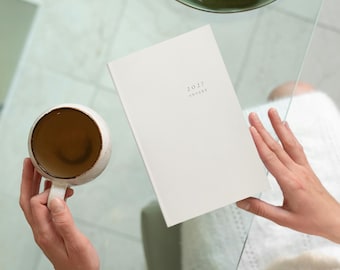 May include: A white rectangular planner with the text 2027 printed on the front cover, positioned next to a round ceramic mug containing a brown liquid. The planner has a clean, solid-colored cover and a simple, minimalist design layout.