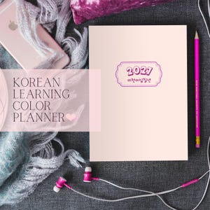 May include: A light pink planner with a rectangular label containing the text 2027 and Korean characters. The planner is positioned next to a pink pencil, a smartphone, and pink wired earbuds on a dark gray textured surface.