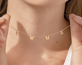 Può includere: Una delicata collana d'oro con le lettere E, M, M e A. Le lettere del ciondolo sono sospese a una sottile catena. La collana è esposta sul collo di una persona, con le mani che tengono delicatamente la catena.