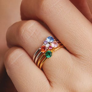 May include: A stack of three thin metal rings worn on a finger. Each ring features a single small round gemstone in a different color, including blue, orange, pink, and green, set in a prong setting. The bands are made of gold and silver tones.