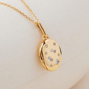 May include: A gold-toned oval locket pendant suspended from a delicate matching chain. The locket features a polished surface adorned with several small, clear, rectangular and round inset stones arranged in a scattered, star-like pattern.