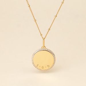 May include: A gold-toned necklace featuring a delicate chain with small beads and a circular pendant. The pendant has a smooth, polished gold center surrounded by a border of small, clear, sparkling stones. The letters S, G, and D are engraved.