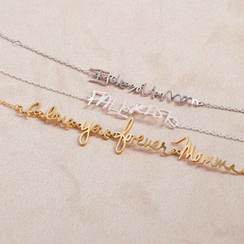 May include: Three delicate chain necklaces featuring cursive metal lettering. The top silver necklace reads I love you mom, the middle silver necklace reads FALL RISK, and the bottom gold necklace reads I love you forever Momma.