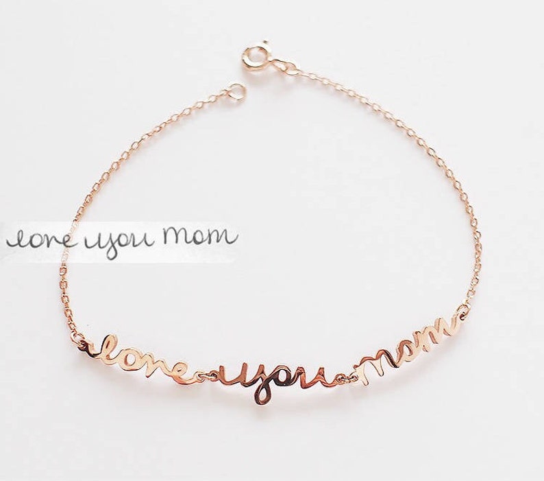 May include: A rose gold-toned metal chain bracelet featuring a script-style pendant that reads "love you mom" in cursive lettering. The delicate link bracelet includes a circular spring ring clasp closure at the top for securing the jewelry piece.