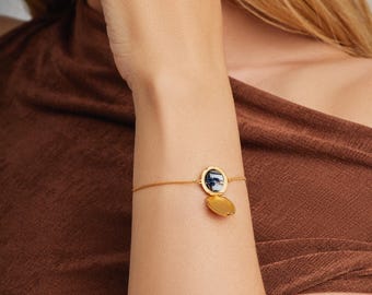 Peut inclure: Un bracelet à chaîne doré doté d'un petit médaillon rond à charnière. Le médaillon est ouvert, révélant un espace intérieur sombre et circulaire, tandis que l'extérieur du médaillon reste fermé et poli. Le bracelet est porté au poignet.