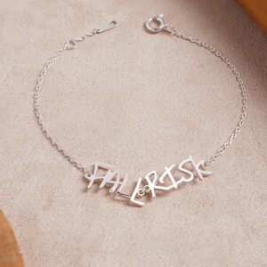 May include: A silver-toned metal chain bracelet featuring a central cutout design that spells out the word "FALL RISK" in a stylized, angular font. The bracelet includes a delicate link chain and a circular spring ring clasp closure.