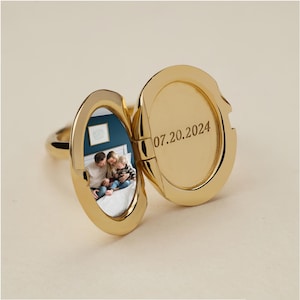 May include: A gold-toned oval locket ring shown in an open position. The left side contains a small photograph of a family sitting on a bed. The right side features the engraved date 07.20.2024 on the smooth, polished interior surface of the metal.