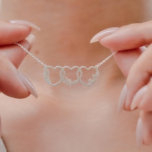 May include: A silver necklace featuring three interconnected heart pendants. Each heart has a name engraved on it, with the word "Love" in the center. The necklace is held up by a person's hands.
