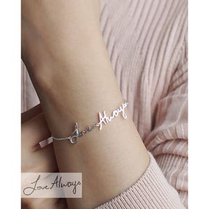 May include: A silver-toned bracelet featuring a delicate chain and a central script metal charm that reads "Love Always". The cursive lettering is integrated directly into the chain design, creating a continuous, fluid aesthetic for the wrist.