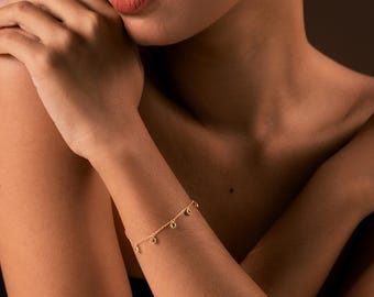 Könnte beinhalten: Ein zartes goldenes Armband mit kleinen, runden Anhängern wird an einem Arm gezeigt. Die Kette des Armbands ist dünn, und die Anhänger scheinen eine hellgrüne Farbe zu haben. Der Hautton ist ein warmer, mittlerer Farbton.