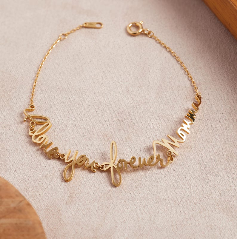 May include: A gold-toned metal bracelet featuring a delicate chain and a central script-style lettering design that reads "love you forever Momma". The piece includes a circular spring ring clasp and a small adjustable chain segment at the end.