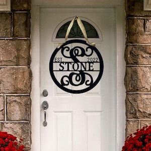 May include: Black metal circular door sign with the name "STONE" and the year "2022". The sign is hanging on a white door with a decorative top. The door is flanked by brick walls and red flowers.