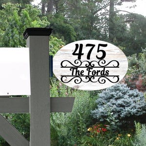 May include: An oval-shaped wooden address sign with the number "475" and the name "The Fords" in black script. The sign is mounted on a gray mailbox post, set against a backdrop of green foliage and a white mailbox.