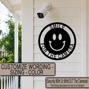 May include: A circular black metal wall sign featuring a cutout smiley face design with the text SMILE FOR THE CAMERA integrated into the border. The sign is mounted on a white horizontal siding wall beneath a white security camera device.