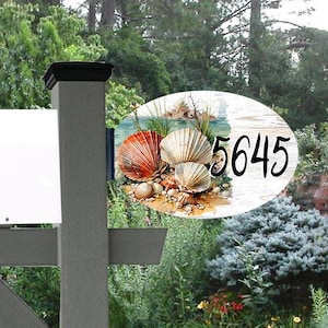 May include: A gray mailbox post with a white mailbox and a decorative address sign. The oval sign features a beach scene with seashells and the number 5645 in black.