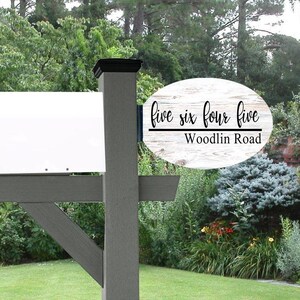 May include: A gray mailbox post with a white mailbox and a sign that reads "Five Six Four Five Woodlin Road" in black script. The sign is oval-shaped with a white wood grain background.