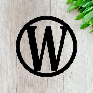 May include: Black wooden letter "W" inside a black circle, set against a light wood grain background. Green leaves are in the upper right corner. The design is simple and modern.