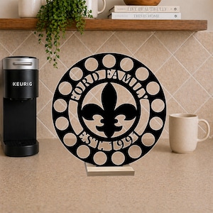 May include: A circular black display stand featuring a fleur-de-lis symbol in the center, surrounded by a ring of circular cutouts. The text "FORD FAMILY" and "EST. 1991" is engraved on the front. The piece rests on a rectangular wooden base.