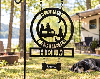 May include: A black metal garden sign hanging from a stand, featuring a circular cutout with a camper, trees, a crescent moon, and a dog. The text reads "HAPPY CAMPERS" and "HELM". A small bone-shaped plaque below is inscribed with "Onyx".