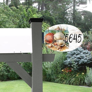 May include: A mailbox post with a decorative address plaque. The oval plaque features a beach scene with seashells and the number 5645 in black. The post is gray with a black top, set against a backdrop of greenery.