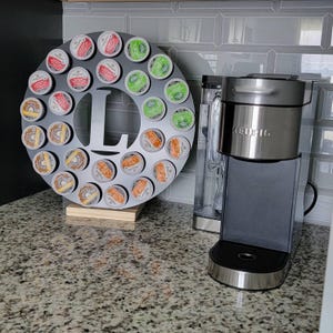 May include: A black circular K-cup holder with the letter "L" cut out, filled with various K-cup pods. A Keurig coffee maker is to the right. The holder sits on a wooden base, and the background is a tiled wall.