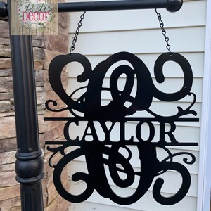 May include: Black metal sign with the name "CAYLOR" and a decorative initial. The sign is suspended by chains from a black metal rod. The sign is mounted on a black post next to a brick wall and white siding.