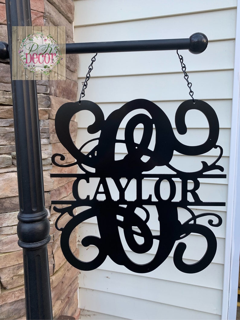 May include: Black metal sign with the name "CAYLOR" and a decorative initial. The sign is suspended by chains from a black metal rod. The sign is mounted on a black post next to a brick wall and white siding.
