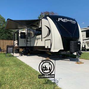 May include: A cream and black Reflection travel trailer with an extended awning is parked on a concrete pad. A black metal sign that says "Happy Campers Welcome" is in the foreground. The sky is blue.
