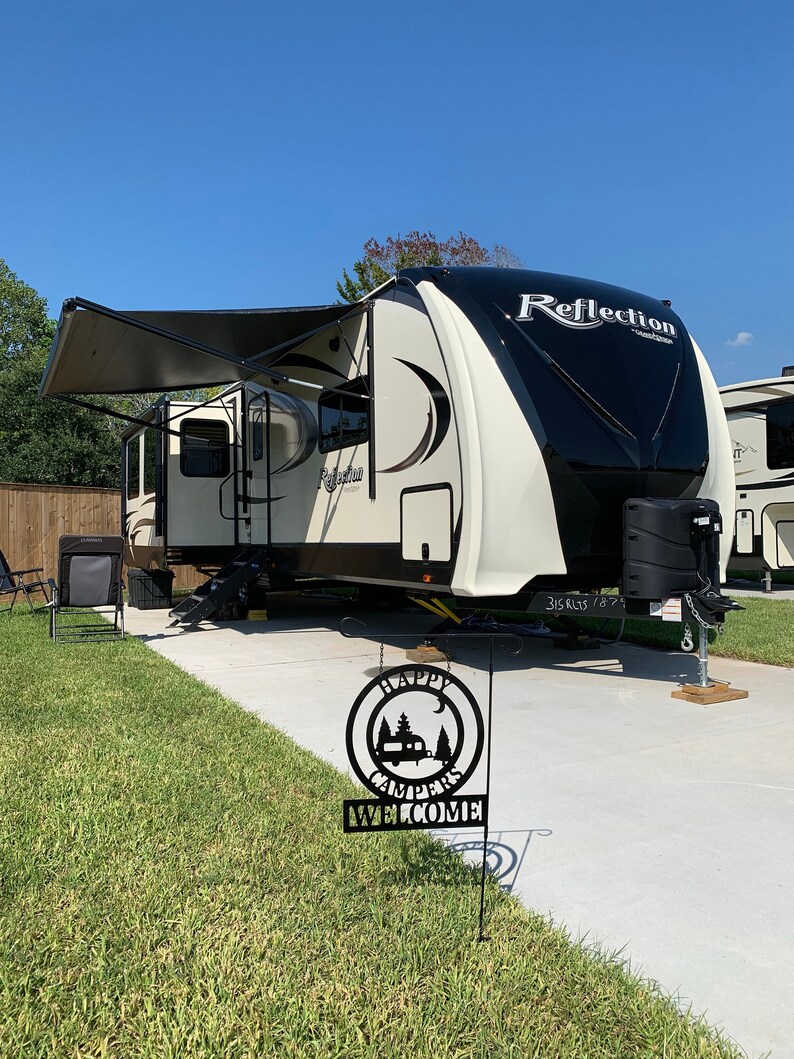 May include: A cream and black Reflection travel trailer with an extended awning is parked on a concrete pad. A black metal sign that says "Happy Campers Welcome" is in the foreground. The sky is blue.