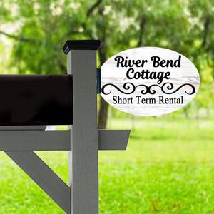 May include: A gray mailbox post with a black top and a white oval sign. The sign reads "River Bend Cottage Short Term Rental" in black script with decorative flourishes. The background is blurred green.