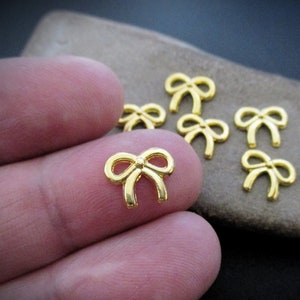 May include: A close-up of eight gold-colored metal bow charms. The bows are small and delicate, with a simple design.