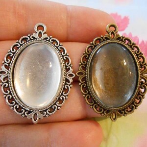 May include: Two silver-tone metal pendant frames with oval glass inserts. One frame has a clear glass insert, and the other has a slightly tinted glass insert.
