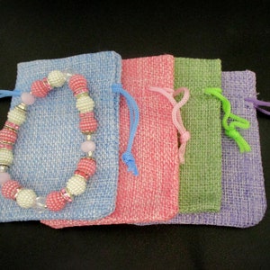 May include: Four small drawstring bags made of burlap fabric in shades of blue, pink, green, and purple. A pink and white beaded bracelet with silver accents is lying on top of the blue bag.