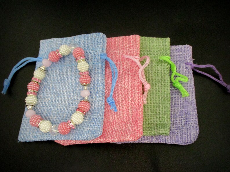 May include: Four small drawstring bags made of burlap fabric in shades of blue, pink, green, and purple. A pink and white beaded bracelet with silver accents is lying on top of the blue bag.