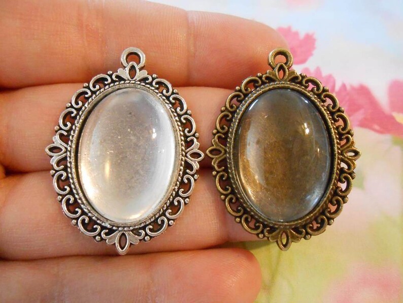 May include: Two silver-tone metal pendant frames with oval glass inserts. One frame has a clear glass insert, and the other has a slightly tinted glass insert.