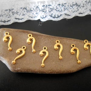 May include: A collection of six gold-toned metal charms shaped like question marks. Each charm features a smooth, polished surface with a small circular loop at the top for attachment, arranged on a flat, irregularly shaped brown stone.