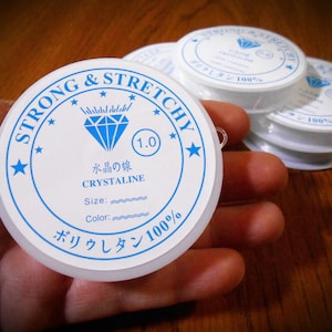 May include: White spools of strong and stretchy crystalline thread, labeled with blue text and a diamond graphic. The text includes "STRONG & STRETCHY", "CRYSTALINE", and "1.0". Several spools are stacked, suggesting a craft supply.