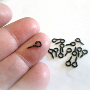 May include: A close-up of a hand holding a black screw-in eyelet. A group of black screw-in eyelets are scattered on a white surface.