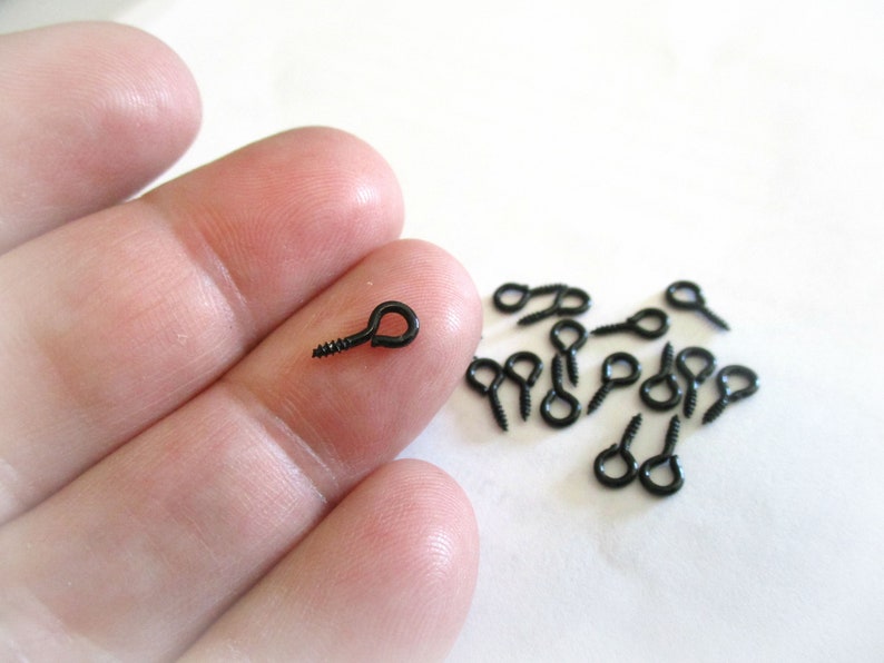 May include: A close-up of a hand holding a black screw-in eyelet. A group of black screw-in eyelets are scattered on a white surface.