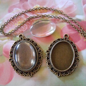 May include: Two oval-shaped pendant settings with ornate bronze frames. One setting has a clear glass insert, the other has a clear plastic insert. A bronze chain is included.