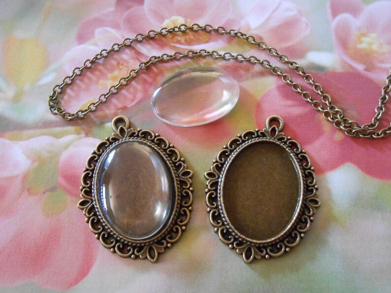 May include: Two oval-shaped pendant settings with ornate bronze frames. One setting has a clear glass insert, the other has a clear plastic insert. A bronze chain is included.