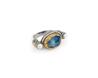 Könnte beinhalten: Ein Silberring mit einem zentralen, oval facettierten blauen Stein in einer goldfarbenen, verzierten Fassung. Die Ringschiene ist beidseitig mit zwei kleinen weißen Perlen besetzt, die in silberne Metallarbeit mit Gravuren eingefasst sind.