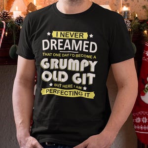Può includere: T-shirt nero con la scritta "I NEVER DREAMED THAT ONE DAY I'D BECOME A GRUMPY OLD GIT BUT HERE I AM PERFECTING IT" in bianco e giallo. La maglietta è girocollo e viene indossata.