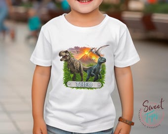 May include: White t-shirt featuring a colorful dinosaur graphic with a volcano backdrop. The design includes a Tyrannosaurus Rex and a Velociraptor, with the name "THEO" on a nameplate. The shirt is a children's garment.