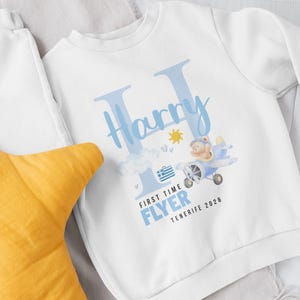 May include: White sweatshirt with a blue design featuring the name "Harry" in a stylized font. A cartoon airplane with a teddy bear pilot is depicted, along with the text "FIRST TIME FLYER TENERIFE 2026". A yellow pillow is in the foreground.
